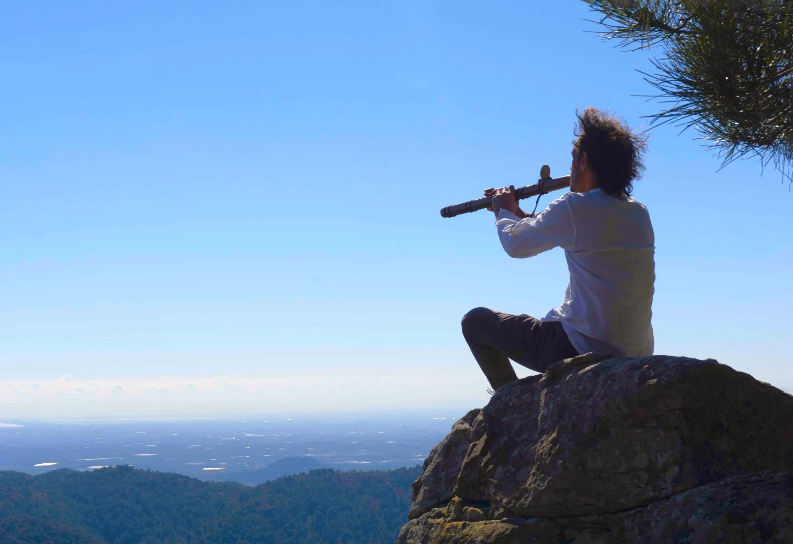 Persona sentada en una roca tocando una flauta con paisaje de montaña y cielo despejado