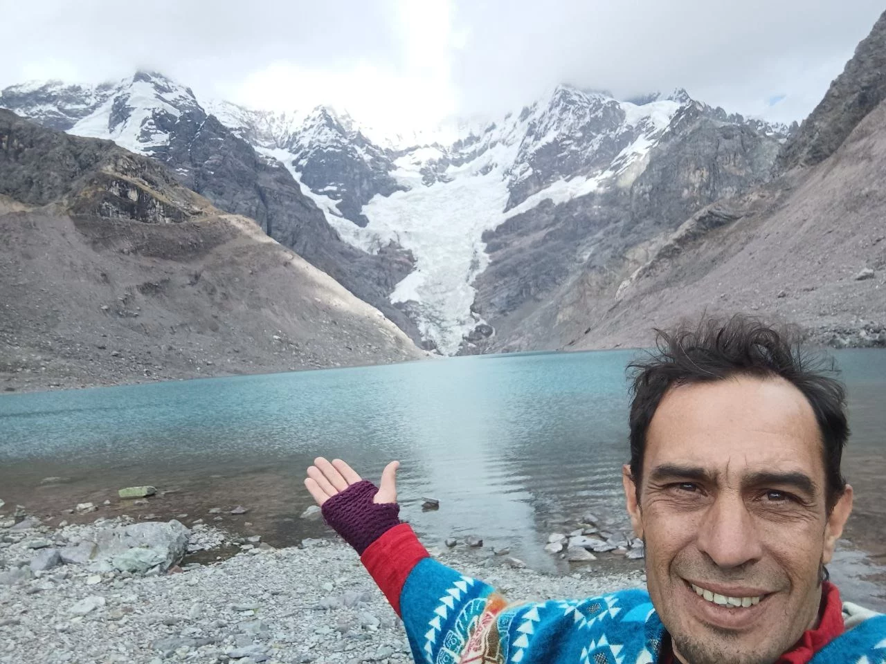 Hombre sonriendo frente a un lago turquesa rodeado de montañas nevadas y rocas